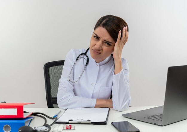 Aching young female doctor wearing medical robe and stethoscope sitting at desk with medical tools and laptop putting hand on head suffering from headache isolated on white wall