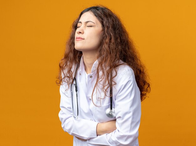 Aching young female doctor wearing medical robe and stethoscope standing in profile view holding belly with closed eyes isolated on orange wall with copy space