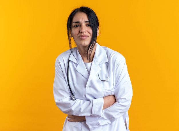 Aching young female doctor wearing medical robe with stethoscope grabbed stomach isolated on yellow wall