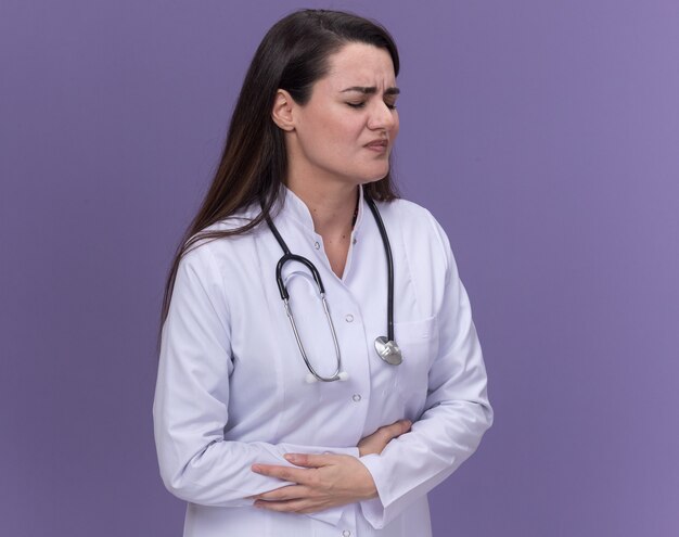 Aching young female doctor wearing medical robe with stethoscope holds belly