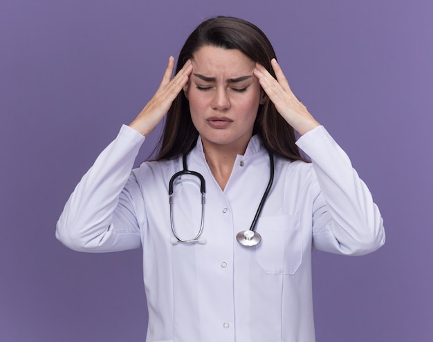 聴診器で医療ローブを身に着けている痛む若い女性医師は、紫色の目を閉じて立っている頭に手を置きます
