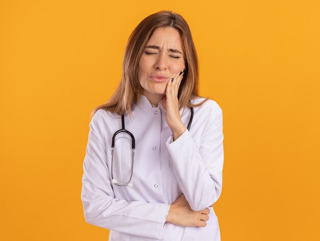 Aching young female doctor wearing medical robe with stethoscope putting hand on aching tooth isolated on yellow wall