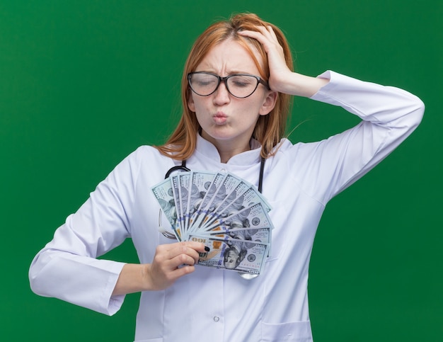 Aching young female ginger doctor wearing medical robe and stethoscope with glasses holding money looking down keeping hand on head having headache isolated on green wall