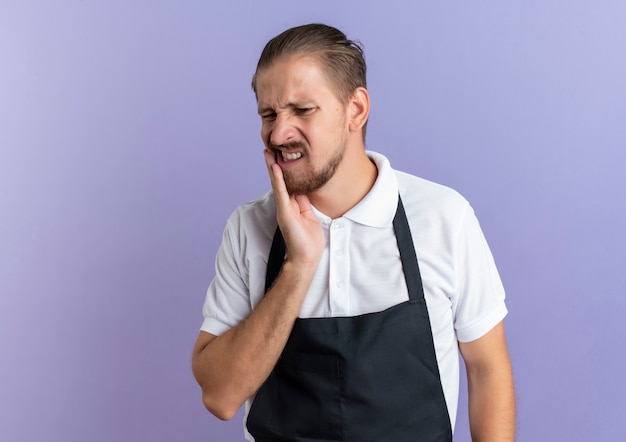 Aching young handsome barber wearing uniform putting hand on cheek suffering from toothache isolated on purple  with copy space