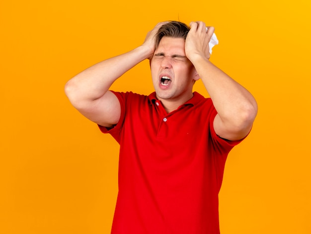 Aching young handsome blonde ill man holding napkin keeping hands on head suffering from headache with closed eyes isolated on orange wall