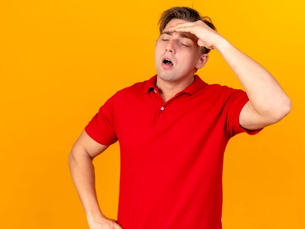 Aching young handsome blonde ill man keeping hand on waist touching forehead having headache with closed eyes isolated on orange background with copy space