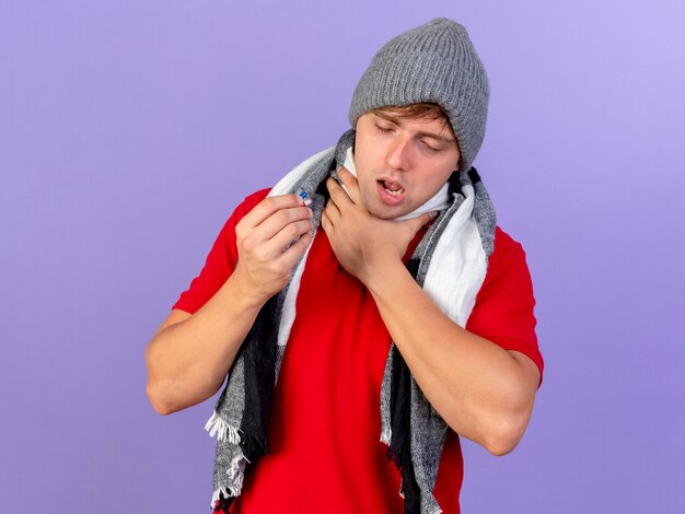 Aching young handsome blonde ill man wearing winter hat and scarf holding medical capsules touching throat with closed eyes isolated on purple background