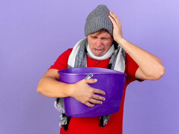 冬の帽子と紫色の壁に分離された頭に触れるプラスチック製のバケツを保持しているスカーフを身に着けている若いハンサムな金髪の病気の男を痛める