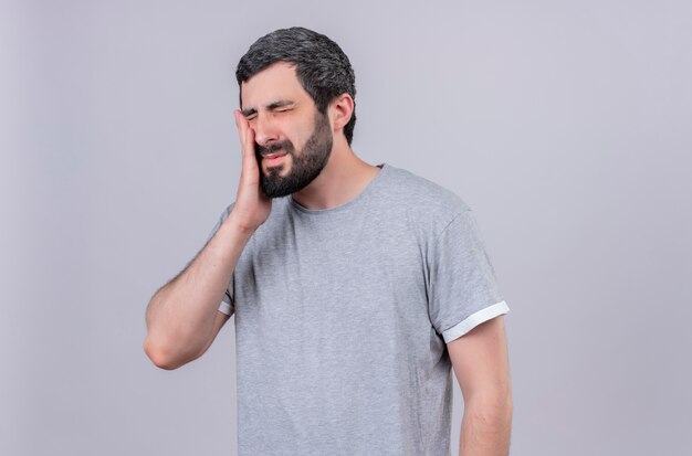 Aching young handsome caucasian man closing his eyes and putting hand on cheek suffering from toothache isolated on white  with copy space