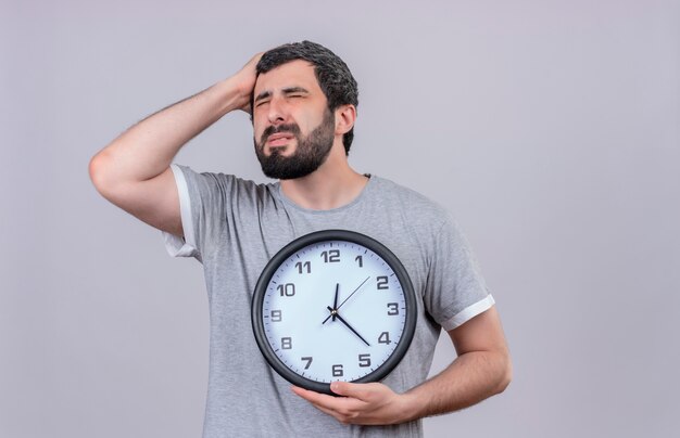 Aching young handsome caucasian man holding clock putting hand on head suffering from pain with closed eyes isolated on white  with copy space