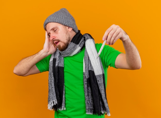 Free photo aching young handsome ill man wearing winter hat and scarf holding thermometer touching head with closed eyes isolated on orange wall