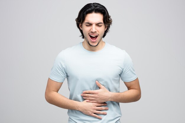 Aching young handsome man keeping hands on belly with closed eyes isolated on white background