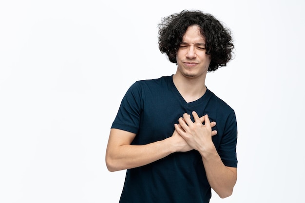 Aching young handsome man keeping hands on chest having heart pain with closed eyes isolated on white background