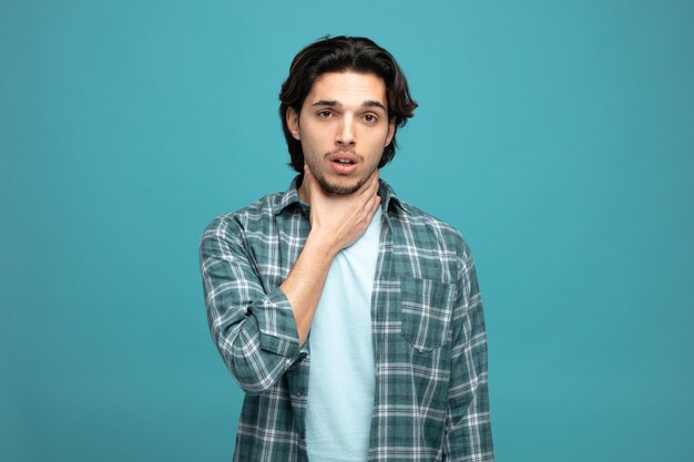 aching young handsome man looking at camera while keeping hand on neck having sore throat isolated on blue background