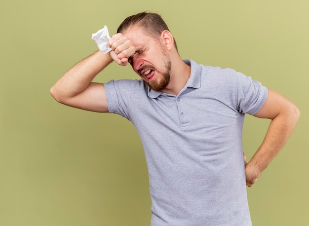 Aching young handsome slavic ill man holding napkin keeping hand on waist and another one on forehead with closed eyes isolated on olive green wall