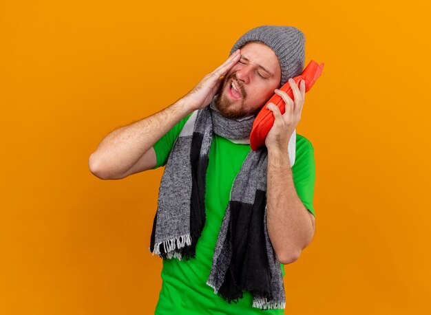 Aching young handsome slavic ill man wearing winter hat and scarf holding hot water bag touching face with it with closed eyes isolated on orange wall with copy space