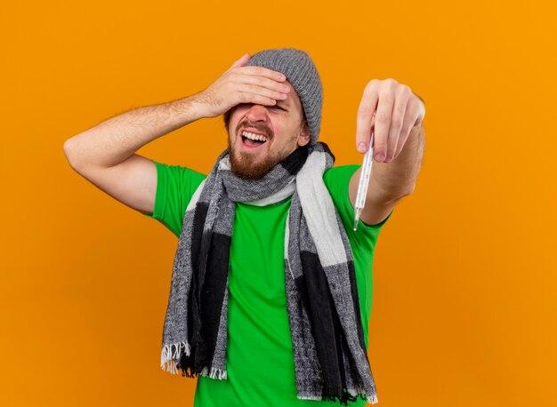 Aching young handsome slavic ill man wearing winter hat and scarf stretching out thermometer  keeping hand on forehead  isolated on orange wall with copy space