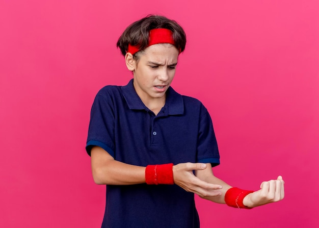 Foto gratuita giovane ragazzo sportivo bello dolorante che indossa la fascia e i braccialetti con le parentesi graffe dentali che osserva e che indica con la mano al suo polso isolato sulla parete rosa con lo spazio della copia