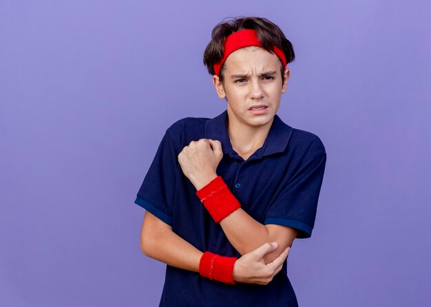 Giovane ragazzo sportivo bello dolorante che indossa fascia e braccialetti con bretelle dentali guardando il gomito della tenuta laterale isolato sulla parete viola