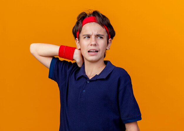 Aching young handsome sporty boy wearing headband and wristbands with dental braces looking at side keeping hand behind neck isolated on orange wall with copy space