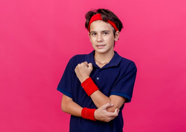 Aching young handsome sporty boy wearing headband and wristbands with dental braces putting hand on elbow looking at camera isolated on crimson background with copy space