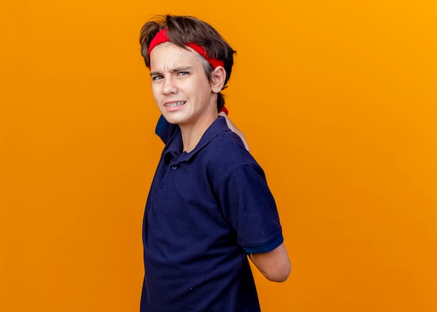 Aching young handsome sporty boy wearing headband and wristbands with dental braces standing in profile view keeping hands behind back isolated on orange wall