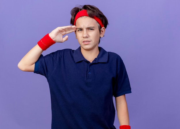 Aching young handsome sporty boy wearing headband and wristbands with dental braces touching head looking at front isolated on purple wall with copy space