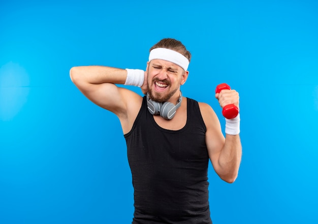 Aching young handsome sporty man wearing headband and wristbands and headphones around neck holding dumbbell and putting hand on neck isolated on blue wall