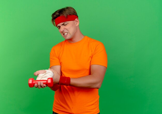 Aching young handsome sporty man wearing headband and wristbands holding dumbbell and his injured wrist wrapped with bandage isolated on green  with copy space