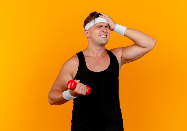 Aching young handsome sporty man wearing headband and wristbands holding dumbbell putting hand on head isolated on orange with copy space