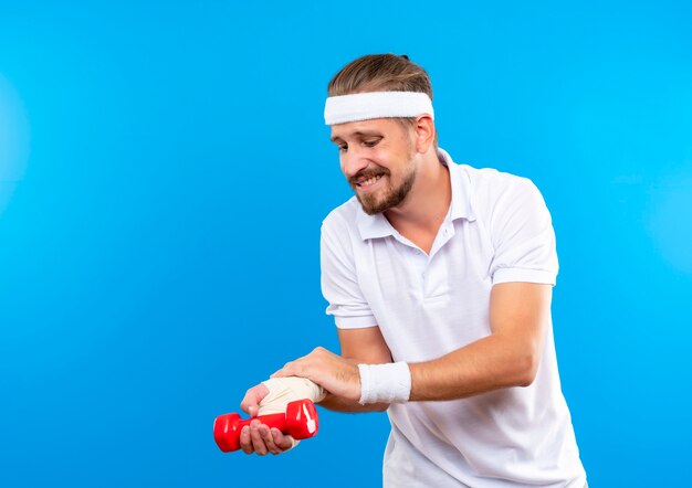 Aching young handsome sporty man wearing headband and wristbands holding dumbbell and putting hand on his injured wrist wrapped with bandage isolated on blue wall with copy space