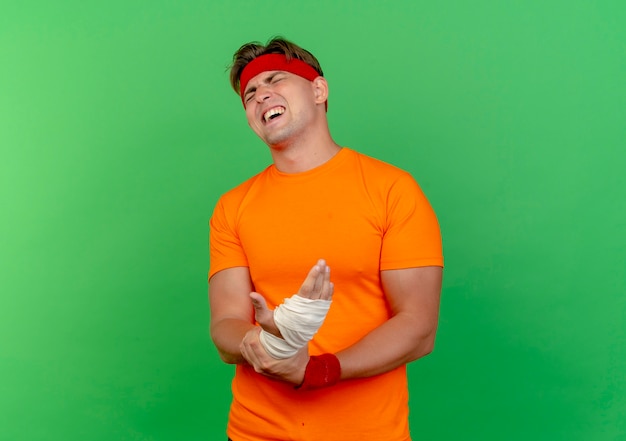 Free photo aching young handsome sporty man wearing headband and wristbands holding his injured wrist wrapped with bandage with closed eyes isolated on green  with copy space