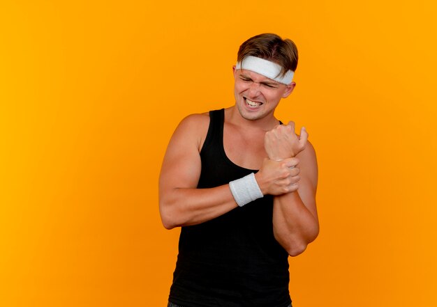 Aching young handsome sporty man wearing headband and wristbands holding wrist isolated on orange  with copy space