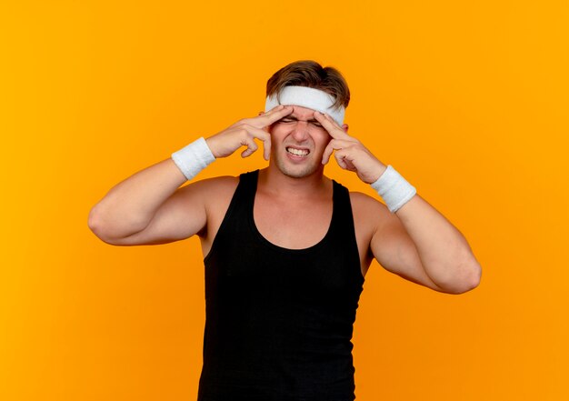 Aching young handsome sporty man wearing headband and wristbands putting fingers on forehead suffering from headache isolated on orange 