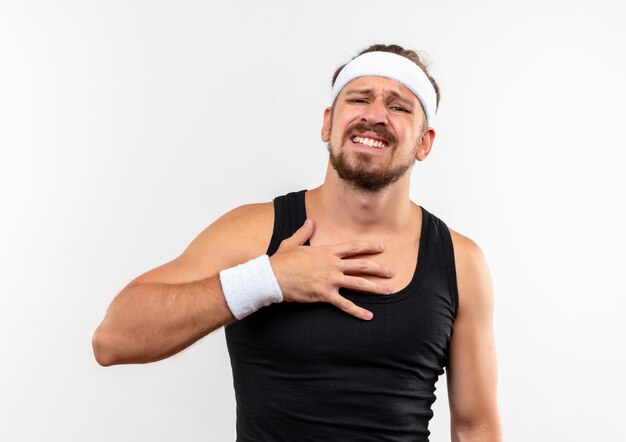 Aching young handsome sporty man wearing headband and wristbands putting hand on chest looking  isolated on white wall