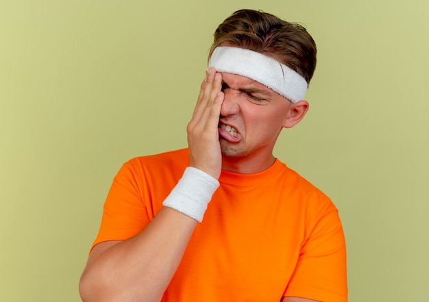 Free photo aching young handsome sporty man wearing headband and wristbands putting hand on eye suffering from pain isolated on olive green