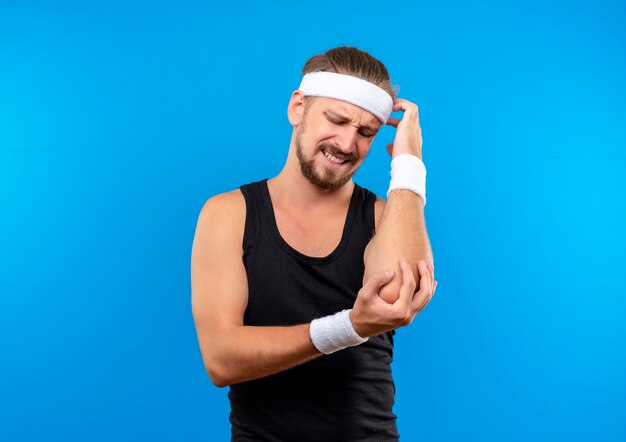 Aching young handsome sporty man wearing headband and wristbands putting hand on his elbow and looking at it isolated on blue wall with copy space