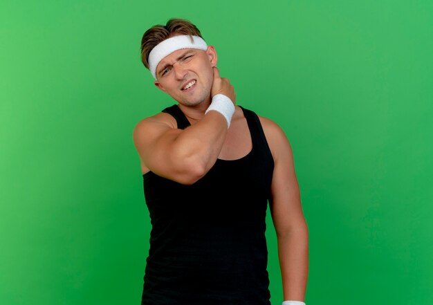 Aching young handsome sporty man wearing headband and wristbands putting hand on neck isolated on green  with copy space