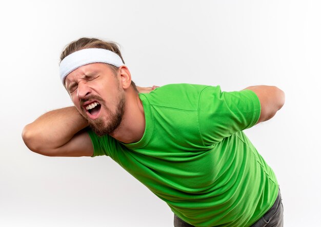 Aching young handsome sporty man wearing headband and wristbands putting hands behind his back with closed eyes isolated on white wall