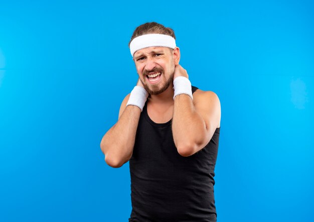 Aching young handsome sporty man wearing headband and wristbands putting hands on his neck isolated on blue wall with copy space