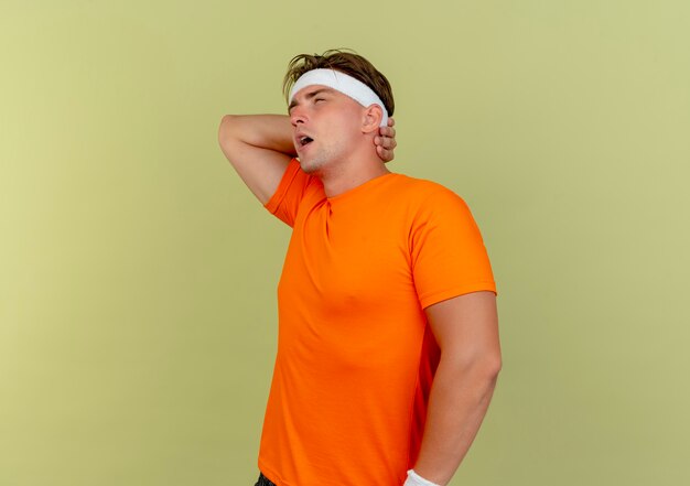 Aching young handsome sporty man wearing headband and wristbands standing in profile view putting hand behind neck looking straight isolated on olive green  with copy space