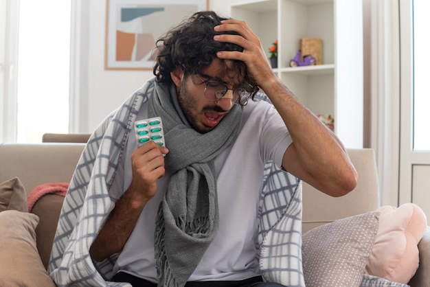 aching young ill caucasian man in optical glasses wrapped in plaid with scarf around his neck putting hand on his head and holding medicine blister pack sitting on couch at living room