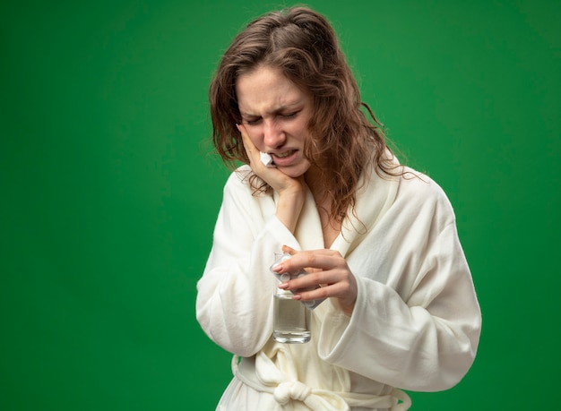 Aching young ill girl wearing white robe holding glass of water putting hand on cheek isolated on green