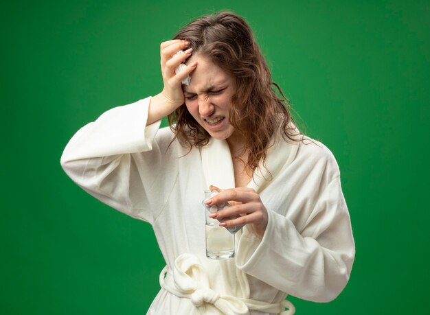 Aching young ill girl wearing white robe holding glass of water putting hand on forehead isolated on green