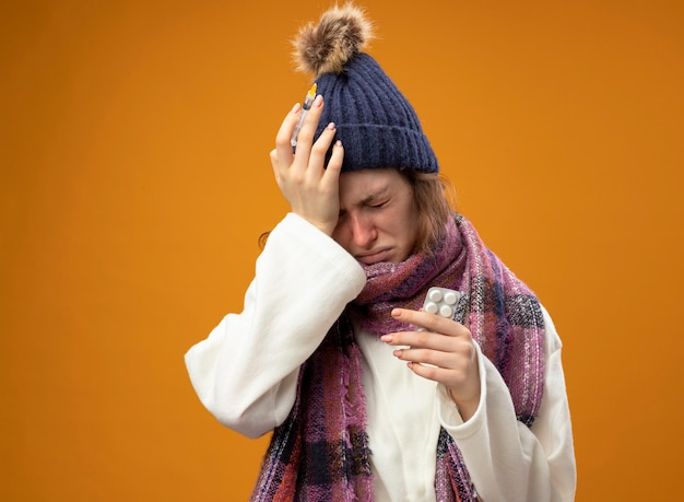 Больная молодая больная девушка в белом халате и зимней шапке с шарфом держит шприц с таблетками, положив руку на лоб, изолированный на оранжевом