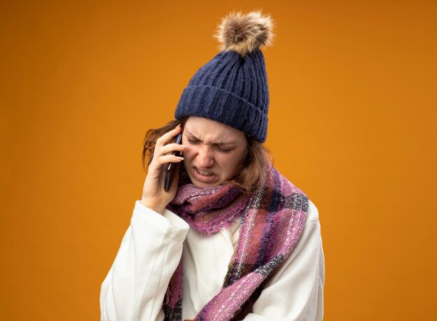 스카프와 흰 가운과 겨울 모자를 쓰고 아프고 아픈 소녀가 오렌지 벽에 고립 된 전화로 말합니다.