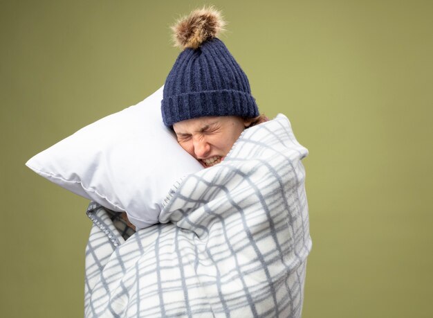 Aching young ill girl wearing white robe and winter hat with scarf wrapped in plaid hugged pillow