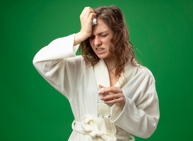 Aching young ill girl with closed eyes wearing white robe holding glass of water and putting hand on forehead isolated on green