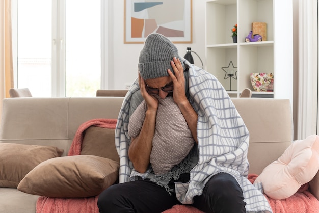 aching young ill man in optical glasses wrapped in plaid with scarf around his neck wearing winter hat putting hands on head holding pillow sitting on couch at living room