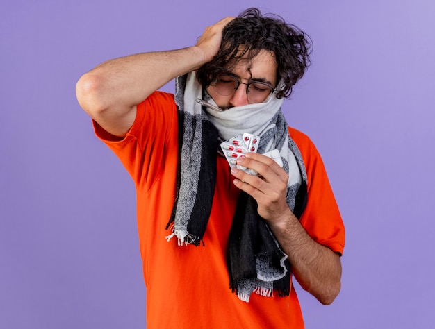 Aching young ill man wearing glasses and scarf putting hand on head holding medical pills with closed eyes isolated on purple wall